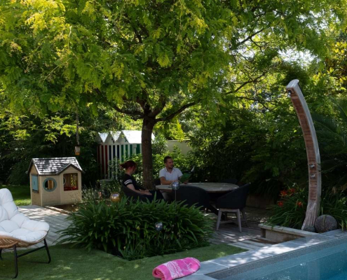 Jardin paysager avec piscine, terrasse en bois et espace de détente ombragé, entouré d’une végétation dense et exotique créant une ambiance conviviale.