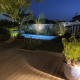 Terrasse en bois avec piscine éclairée et jardin paysager de nuit, mise en valeur par un éclairage extérieur, réalisation par Au Jardin des Rêves.