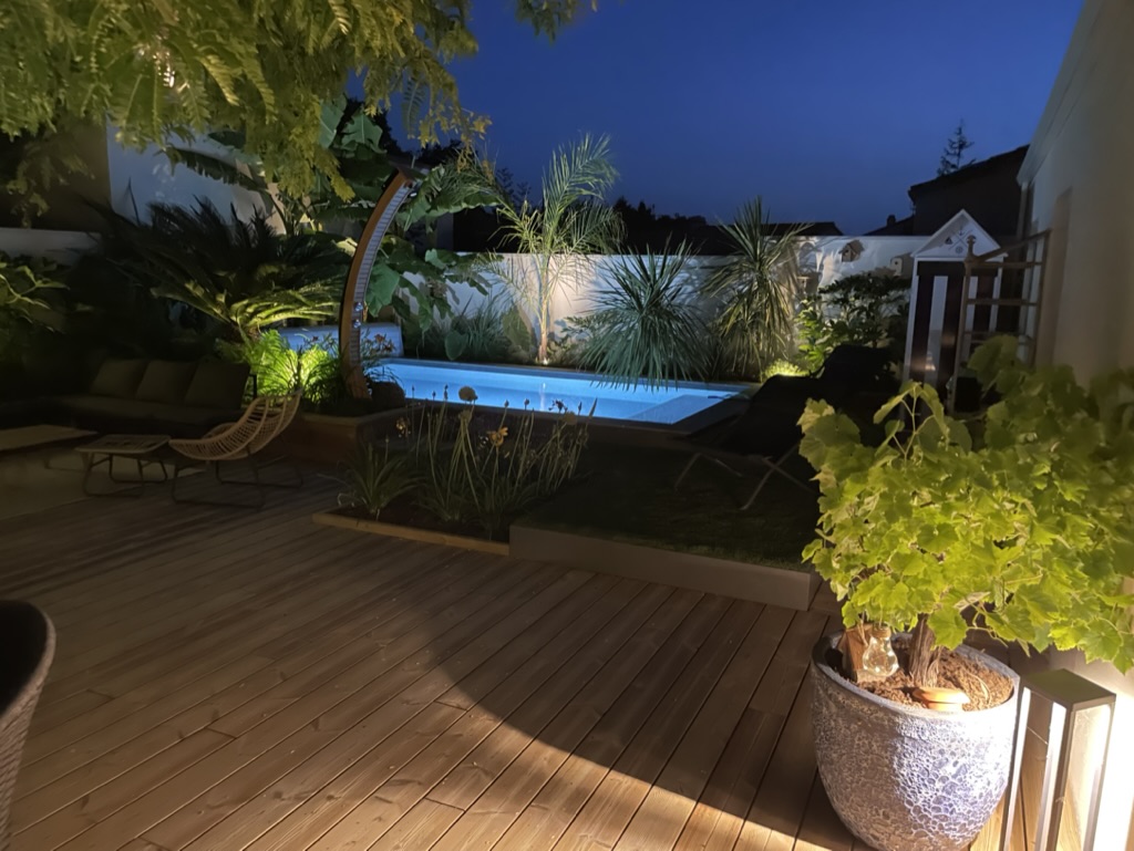 Terrasse en bois avec piscine éclairée et jardin paysager de nuit, mise en valeur par un éclairage extérieur, réalisation par Au Jardin des Rêves.