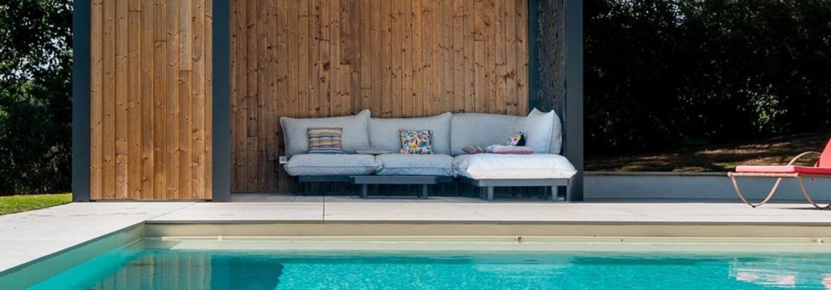 Piscine moderne avec pool house en bois, salon extérieur couvert et terrasse claire dans un jardin aménagé