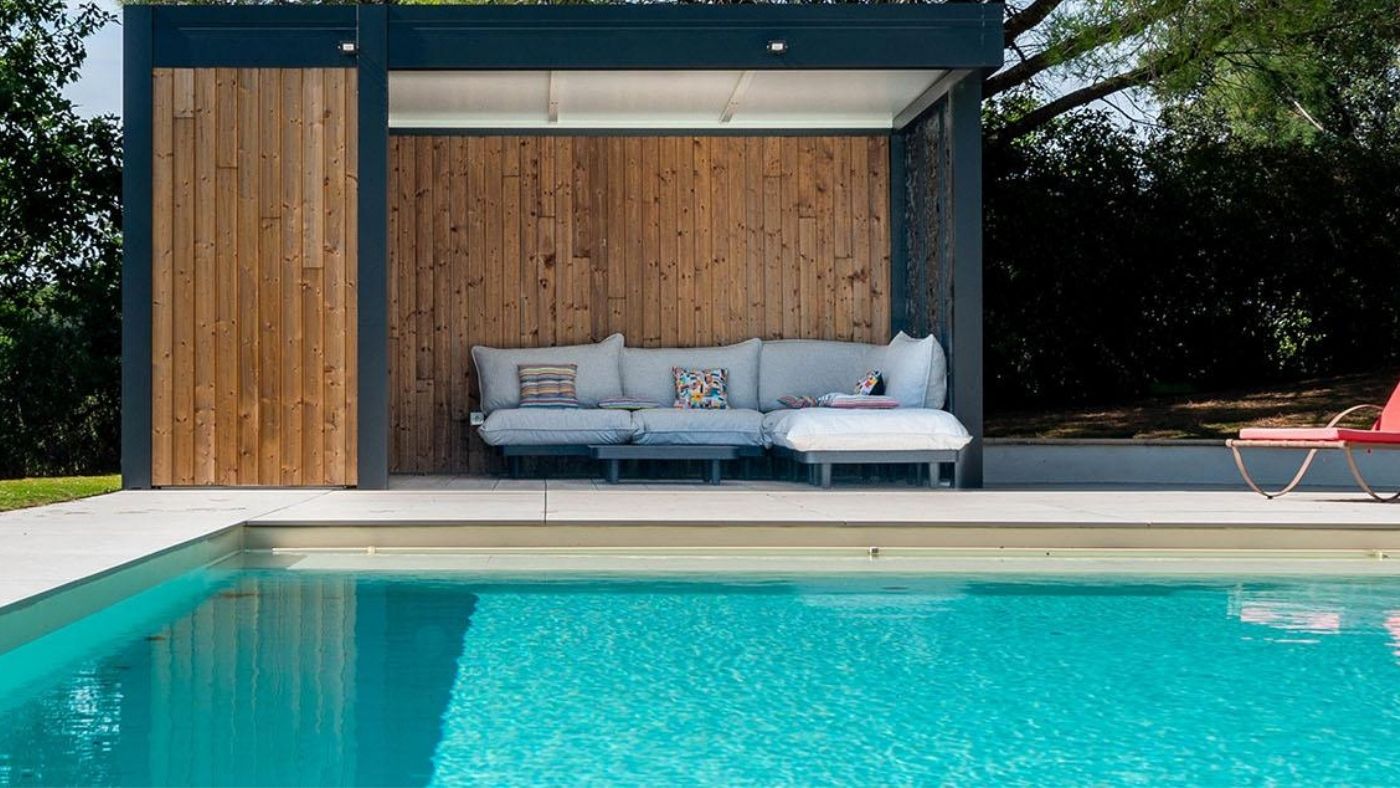 Piscine moderne avec pool house en bois, salon extérieur couvert et terrasse claire dans un jardin aménagé